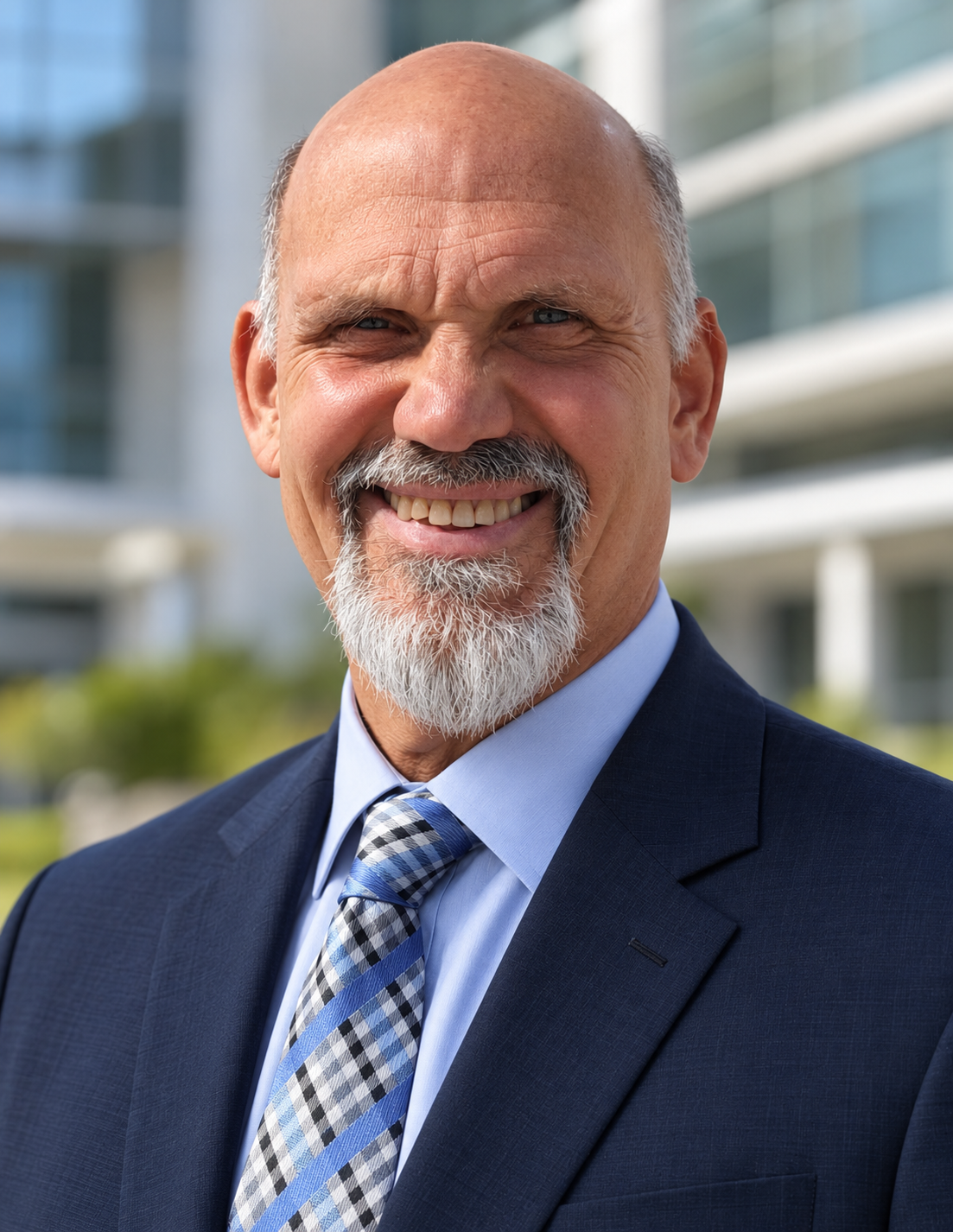 Portrait of Dr. Russ L'HommeDieu, smiling, in a navy suit and blue plaid tie outdoors in front of a modern glass building.